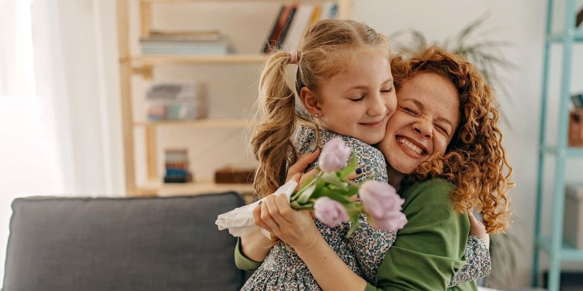 Moeder knuffelt met haar dochter terwijl ze zitten op de bank. De moeder met groene trui heeft een bos tulpen in haar hand.