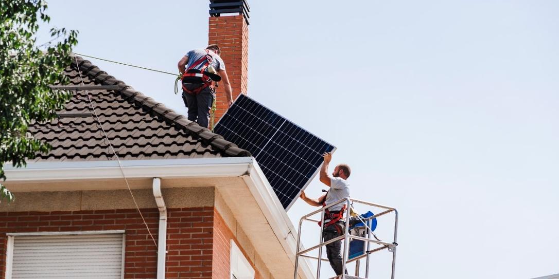 Twee installateurs staan op het dak van een huis en op de hoogwerker met een zonnepaneel in hun handen