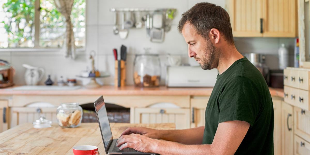Man werkt aan keukentafel op zijn laptop en kijkt serieus richting het scherm.