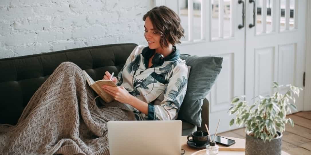 Vrouw zit op de bank onder een deken te schrijven in een boekje met haar koptelefoon op en een laptop en telefoon op de tafel naast haar