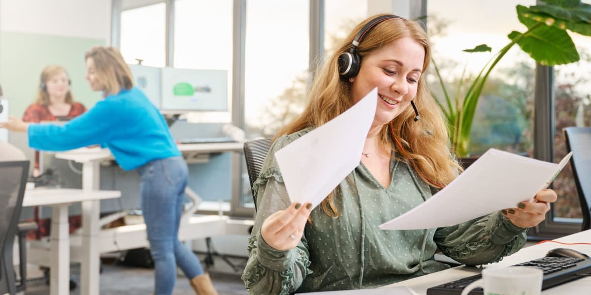 Ervaren jurist met headset kijkt lachend naar haar documenten, collega's op de achtergrond